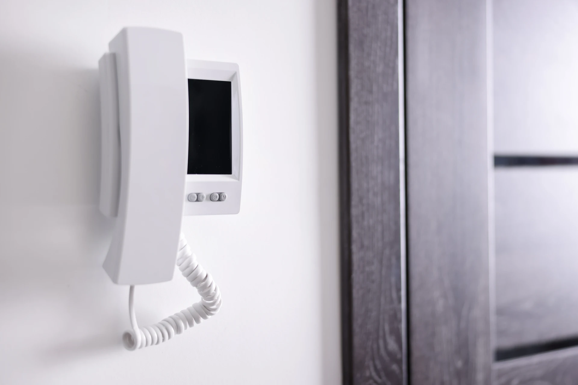 Interphone vidéo couleur à écran tactile IPS monté sur le montant de porte d'entrée d'une maison résidentielle. Finition inox brossé, bouton d'appel lumineux LED, électricien en uniforme installant et testant l'appareil, gros plan sur les connexions électriques au-dessus du cadre de porte avec gaine électrique grise visible.
